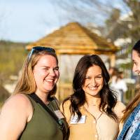Three young women outside at event, standing together and smiling with the sun shining on them